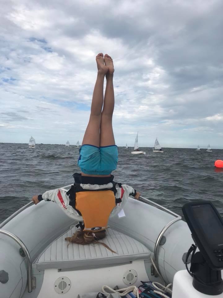 A person in a life jacket and blue shorts performs a headstand on the bow of an inflatable boat, with sailboats and choppy water in the background under a cloudy sky.