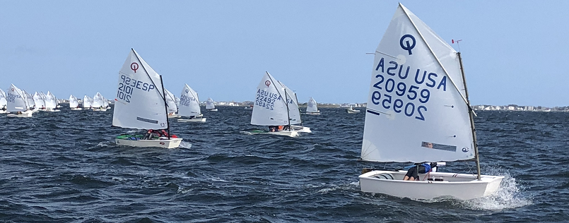 Children sail small white dinghies with numbers on the sails across choppy blue water in a sailboat race; more boats can be seen in the background under a clear sky.