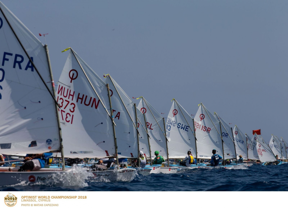 A row of young sailors in small sailboats compete in a regatta, each with different country codes on their sails, during the Optimist World Championship 2018 in Limassol, Cyprus.