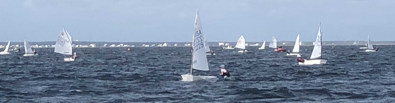 Several small sailboats with solo sailors are spread out on a choppy body of water under a cloudy sky, suggesting a sailing event or regatta. The shoreline is visible in the distance.