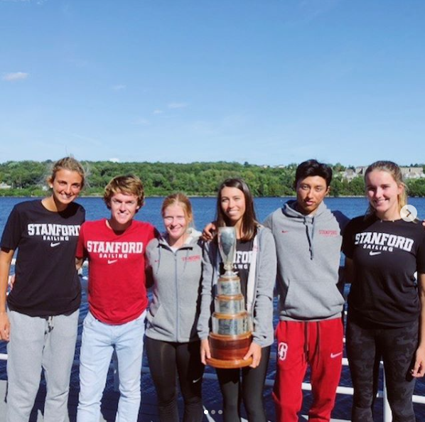 Six young adults stand outdoors by a lake, smiling and posing together. They wear Stanford Sailing attire and one holds a large trophy. The background features water, trees, and a clear blue sky.