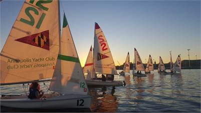Several small sailboats with colorful sails, marked "Annapolis Yacht Club," float on calm water at sunset. The sky is clear, and people are visible in the boats, preparing to sail or racing.