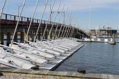 A row of small sailboats with their masts up are docked along a wooden pier beside a bridge over calm water on a clear, sunny day.
