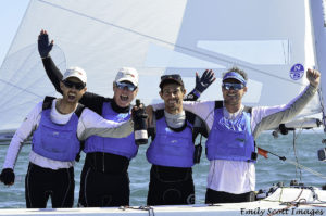Four smiling men wearing blue life vests and sunglasses stand on a sailboat, celebrating with arms around each other and holding a bottle. The sea and sail are visible in the background.
