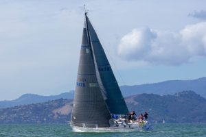 A white sailboat with a large dark sail glides across the water, crew members on board. Hills and a partly cloudy sky are visible in the background.