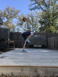 A woman in athletic clothing exercises outdoors on a wooden deck, lifting one knee high. Trees with autumn leaves and a wooden fence surround the area. The sky is clear and sunny.