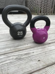 Two kettlebells on a wooden deck: one is gray and labeled “25,” and the other is smaller, purple, and labeled “12.” A fence and outdoor furniture are in the background.