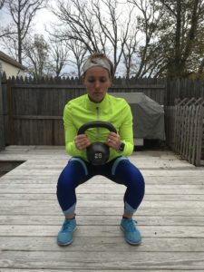 A woman in athletic clothing squats on a wooden deck while holding a 25-pound kettlebell with both hands. She wears a neon yellow jacket, blue leggings, blue shoes, and a headband. Trees and fences are in the background.
