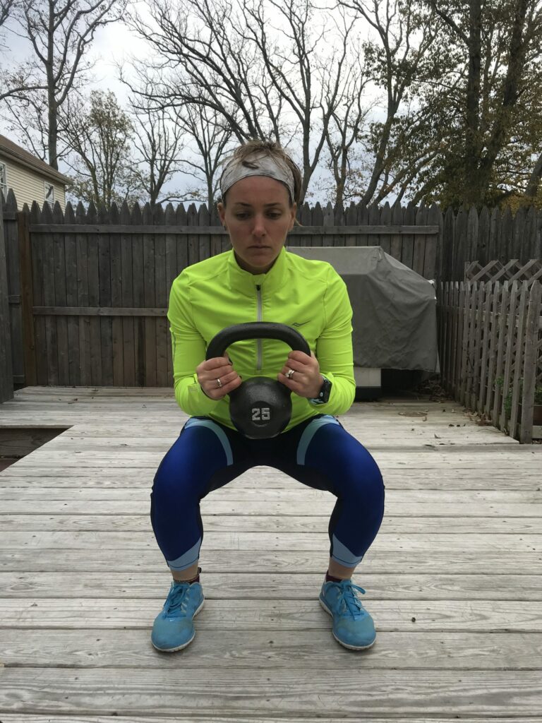 A woman in athletic clothing squats on a wooden deck while holding a 25-pound kettlebell with both hands. She wears a neon yellow jacket, blue leggings, blue shoes, and a headband. Trees and fences are in the background.