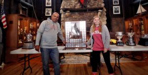 Two people stand in a lodge-like room, smiling and posing with a large sailboat trophy on a table. The room features wood walls, trophy cases, a stone fireplace, and framed photos on the wall behind them.
