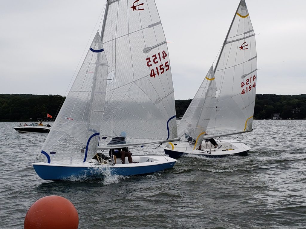 Two small sailboats with white sails race closely on a lake, creating splashes. The boats have the numbers '4155' and '4154.' In the background are trees, houses, and a distant motorboat. An orange buoy floats in the foreground.