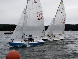 Two small sailboats with white sails race closely on a lake, creating splashes. The boats have the numbers "4155" and "4154." In the background are trees, houses, and a distant motorboat. An orange buoy floats in the foreground.