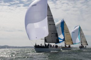 Three sailboats with large, billowing sails race side by side on the water under a partly cloudy sky, with distant land visible in the background.