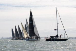 Several sailboats with tall sails are lined up and racing on a body of water under a cloudy sky. One boat with a flag is in the foreground, while others are behind it, forming a diagonal line. A bridge is visible in the distance.