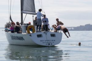 A group of people relax on a sailboat named "MUDSHARK" in calm water. One person dives off the back of the boat while another swims nearby. The background includes a distant shoreline and a bridge.