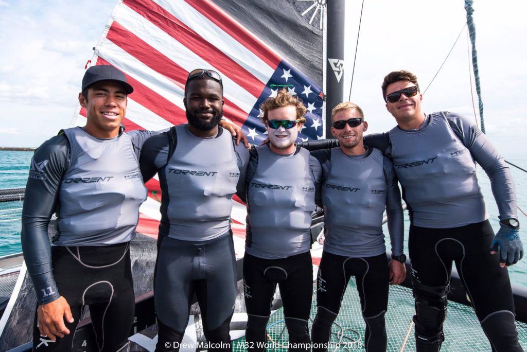 Five men wearing gray 'TORRENT' shirts and black wetsuits stand smiling on a boat with an American flag in the background. They appear to be part of a sailing team, with blue water and sky behind them.