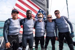 Five men wearing gray "TORRENT" shirts and black wetsuits stand smiling on a boat with an American flag in the background. They appear to be part of a sailing team, with blue water and sky behind them.