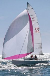 A small sailboat with two people aboard sails on a sunny day. The boat has a large white and pink sail and glides over shimmering water under a clear blue sky.