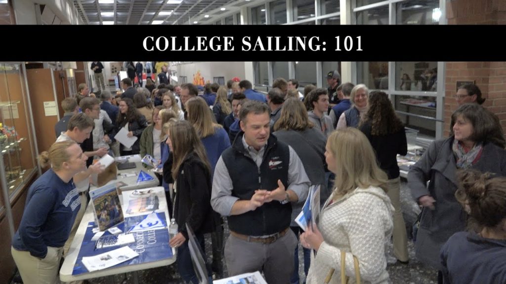A busy indoor event with many people engaging at tables displaying college sailing materials, under a banner reading 'College Sailing: 101.' The setting appears to be a hallway or lobby with large windows.