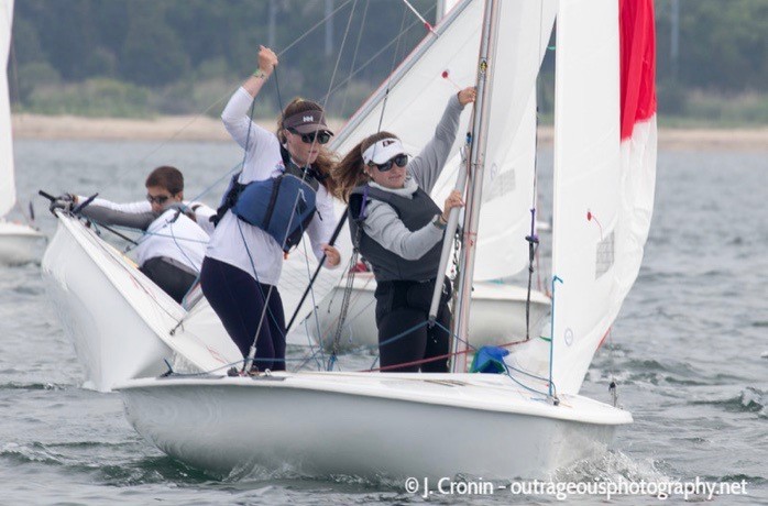 Two women wearing life jackets and sunglasses sail a small boat in a race, with another boat and sailors visible behind them on the water. The shoreline and trees are in the background.