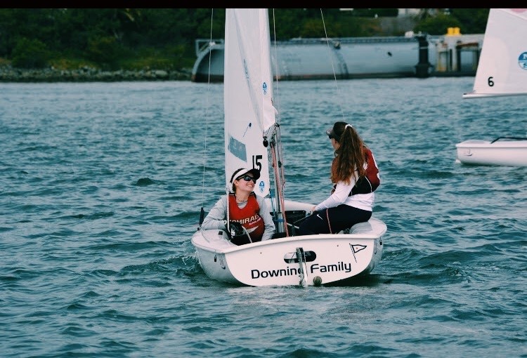 Two people are sailing a small white dinghy labeled 'Downing Family' on the water, both wearing life jackets. One is sitting facing forward, while the other faces them. Trees and a dock are visible in the background.