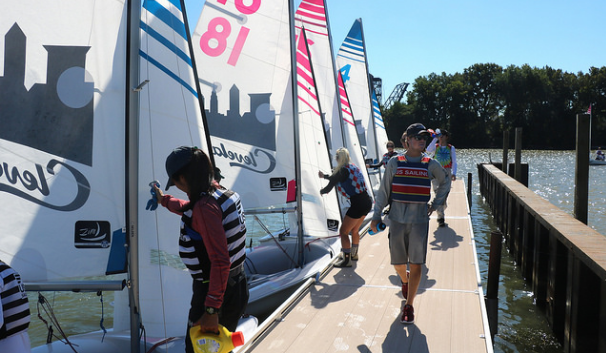 People in life jackets prepare sailboats at a dock on a sunny day. Some adjust sails while others stand or walk on the dock near the water, with trees and buildings visible in the background.
