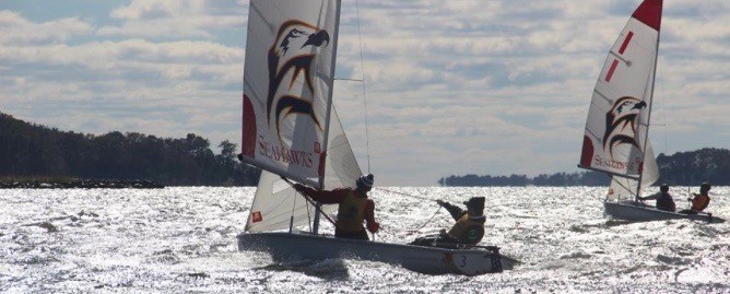 Two sailboats with eagle designs on their sails race on a sunlit, choppy body of water. Three people are visible, wearing life jackets and cold weather gear, with trees and distant land in the background.