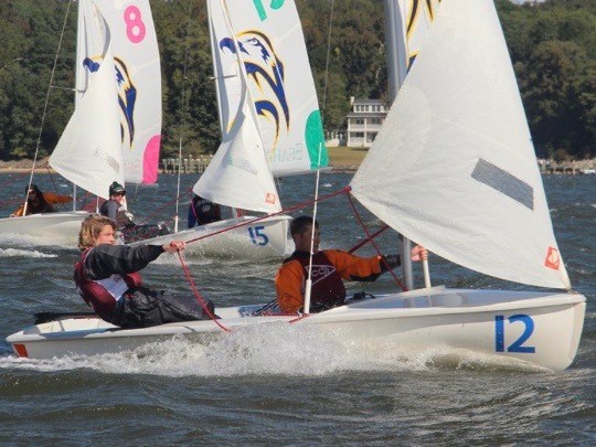 Two sailboats with teams of two people each compete on a lake, creating splashes as they move. The front boat is numbered 12, and trees and a white house are visible on the shore in the background.