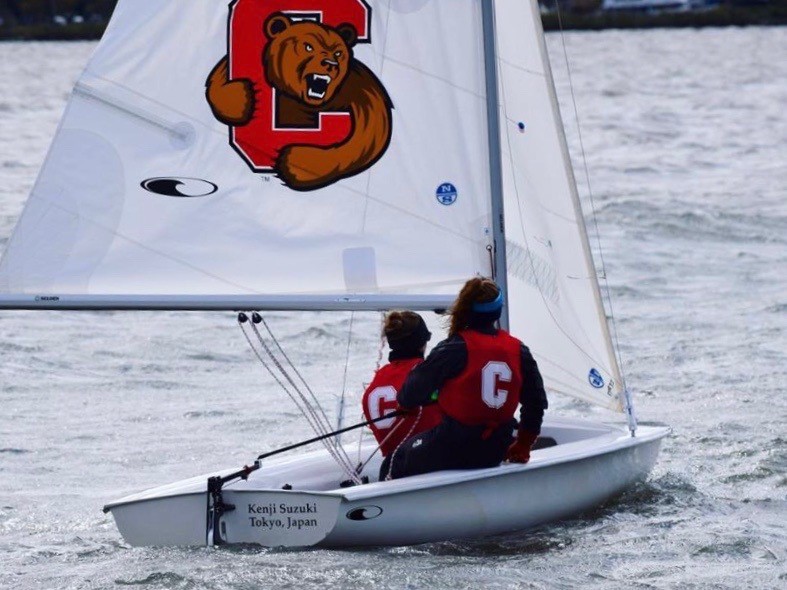 Two people wearing life vests with a large "C" sail a small white boat on choppy water. The sail features a roaring bear and red "C" logo. A nameplate reads "Kenji Suzuki, Tokyo, Japan.