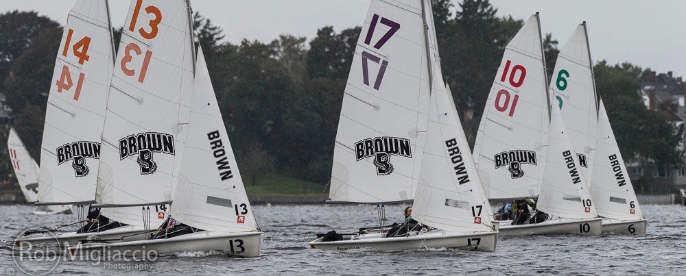 Four Brown University sailboats with numbered sails (13, 17, 10, 6) race on the water near a tree-lined shore, under cloudy skies. The boats have 'BROWN' logos on their sails.