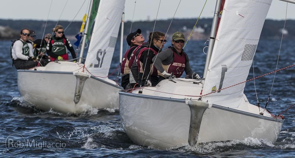Two sailboats with college sailing teams race closely on open water, with crew members adjusting lines and leaning out. The crews wear life jackets and team gear, and land is visible in the distant background.