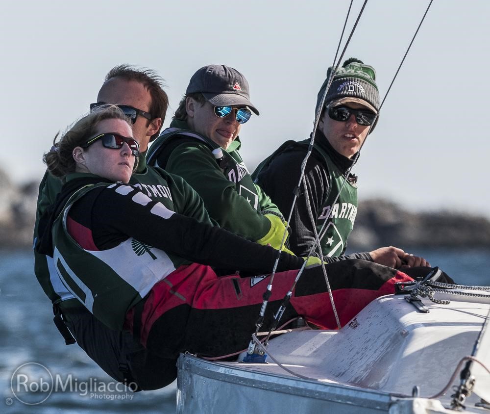 Four people in sailing gear lean out from the side of a white sailboat on the water, concentrating as they navigate. They wear sunglasses and green Dartmouth sweatshirts, with the shoreline blurred in the background.