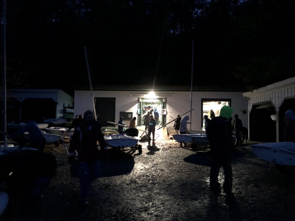 People stand outside a brightly lit building at night, surrounded by small sailboats on trailers. The ground is wet and the area is dimly lit, with shadows and silhouettes clearly visible.