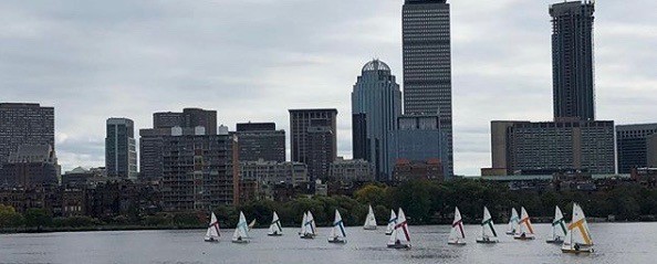 Small sailboats with colorful sails race on a river in front of a city skyline with tall buildings under a cloudy sky.