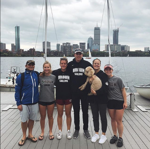 Six people and a dog stand on a dock in front of sailboats, with a city skyline and tall buildings in the background. Four people wear "Brown Sailing" shirts, and the sky is overcast.