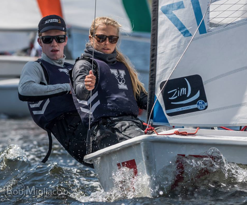 Two young sailors wearing sunglasses and life jackets lean out of a small sailboat as they race on choppy water, focused on guiding the vessel. Other colorful sailboats are visible in the background.