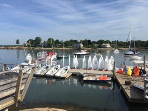 A marina with small sailboats docked at piers, several larger sailboats anchored in calm water, and people preparing boats under a blue sky with trees and houses in the background.
