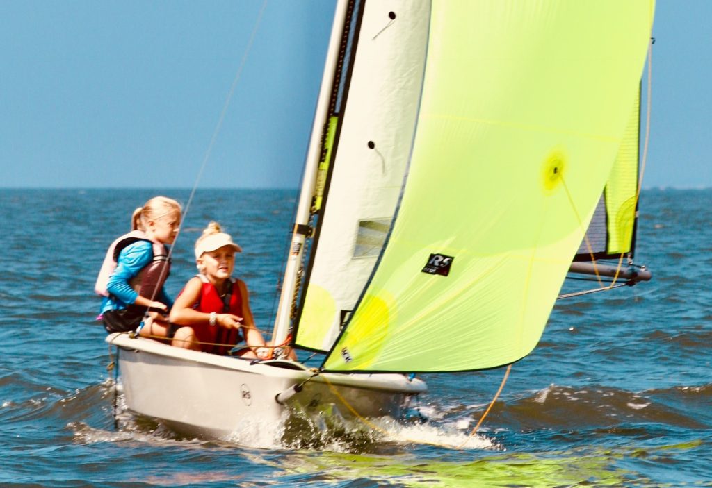 Two children wearing life vests and smiling as they sail a small boat with a bright yellow sail on a sunny day, with blue water and sky in the background.