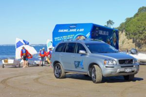 A silver Volvo SUV is parked on a sandy beach near a blue tent and small sailboats. Several people in life jackets and caps stand nearby, preparing for a sailing activity by the water, with trees in the background.