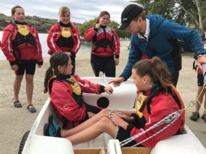 A group of girls wearing red life jackets listen to an instructor by a small boat on shore. Two girls sit inside the boat while the others stand nearby, with water and trees in the background.