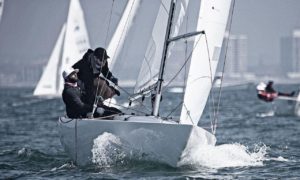 Two people in dark clothing sail a small white sailboat through choppy water, leaning to adjust the sail. Other sailboats and city buildings are visible in the background on a hazy day.