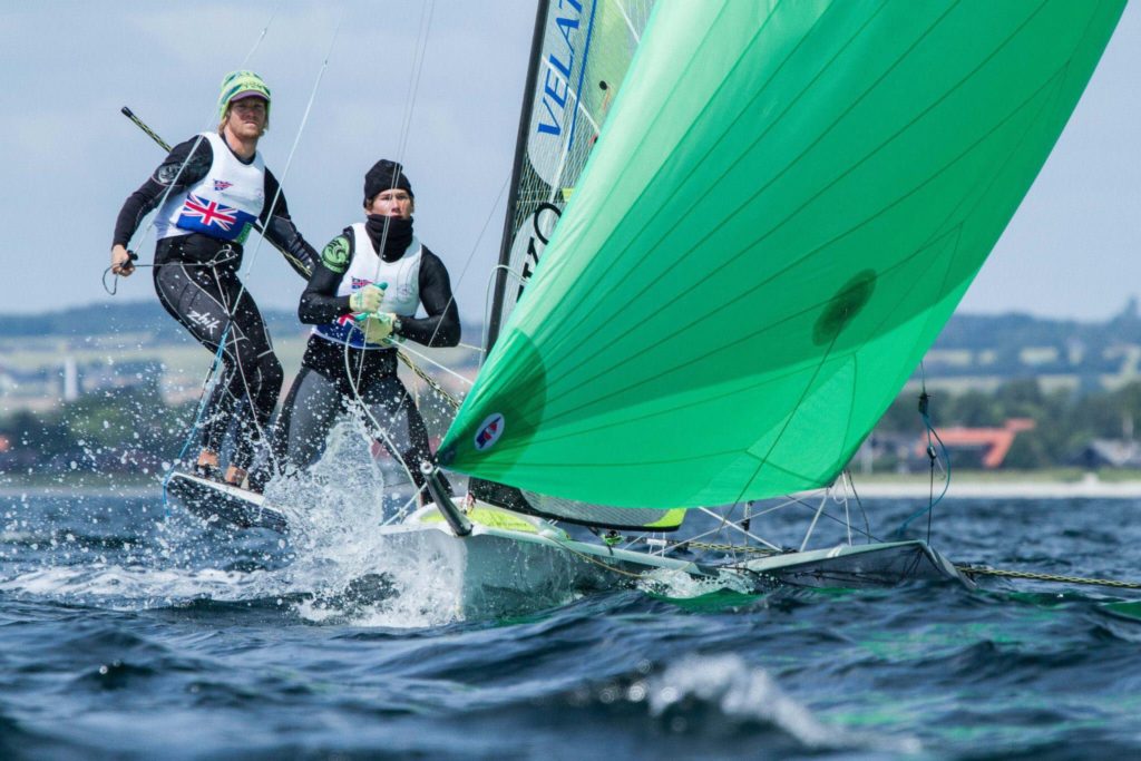Two sailors in wetsuits compete in a sailboat race, riding over choppy water. The boat has a large, bright green sail, and the background shows a shoreline with trees and buildings.