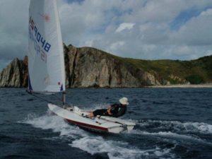 A person in a small sailboat navigates choppy blue water near a rocky, green coastline under a cloudy sky.