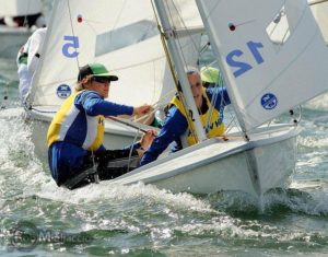 Two sailors wearing life jackets and sunglasses navigate a small sailboat numbered 12 through choppy water during a race, with other boats and crew visible nearby in the background.