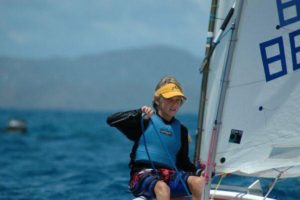 A young person wearing a yellow visor and blue life jacket sails a small boat on the ocean, with mountains visible in the background under a partly cloudy sky.