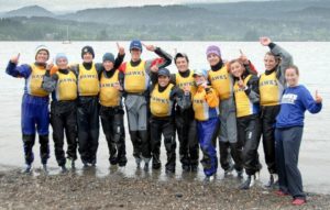 A group of people in yellow life jackets labeled “HAWKS” smile and pose at the shore of a lake, holding up one finger each. Mountains and trees are visible in the background under an overcast sky.