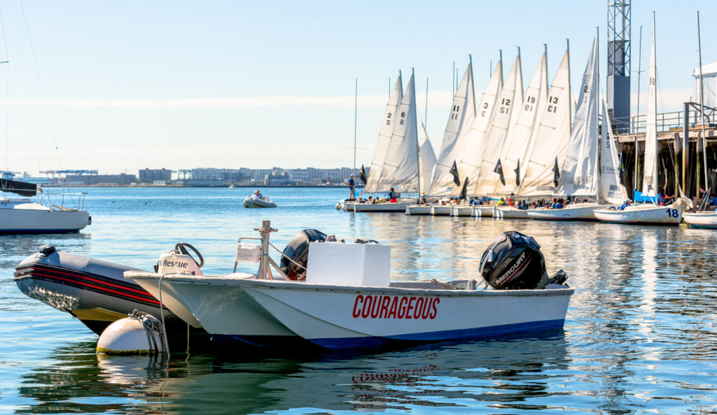 Small motorboat labeled 'Courageous' floats on calm water near a dock with several white sailboats lined up, sails raised. People are gathered on and around the boats, under a clear blue sky.