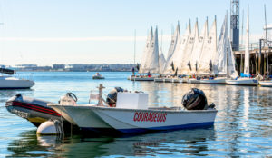 Small motorboat labeled "Courageous" floats on calm water near a dock with several white sailboats lined up, sails raised. People are gathered on and around the boats, under a clear blue sky.