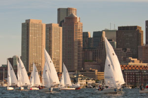 Sailboats racing on the water with their white sails up, set against a city skyline of tall modern buildings in the background under a clear sky.