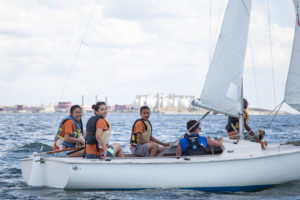 Five young adults wearing life vests smile while sailing a small white sailboat on a calm body of water, with an industrial shoreline and cloudy sky in the background.